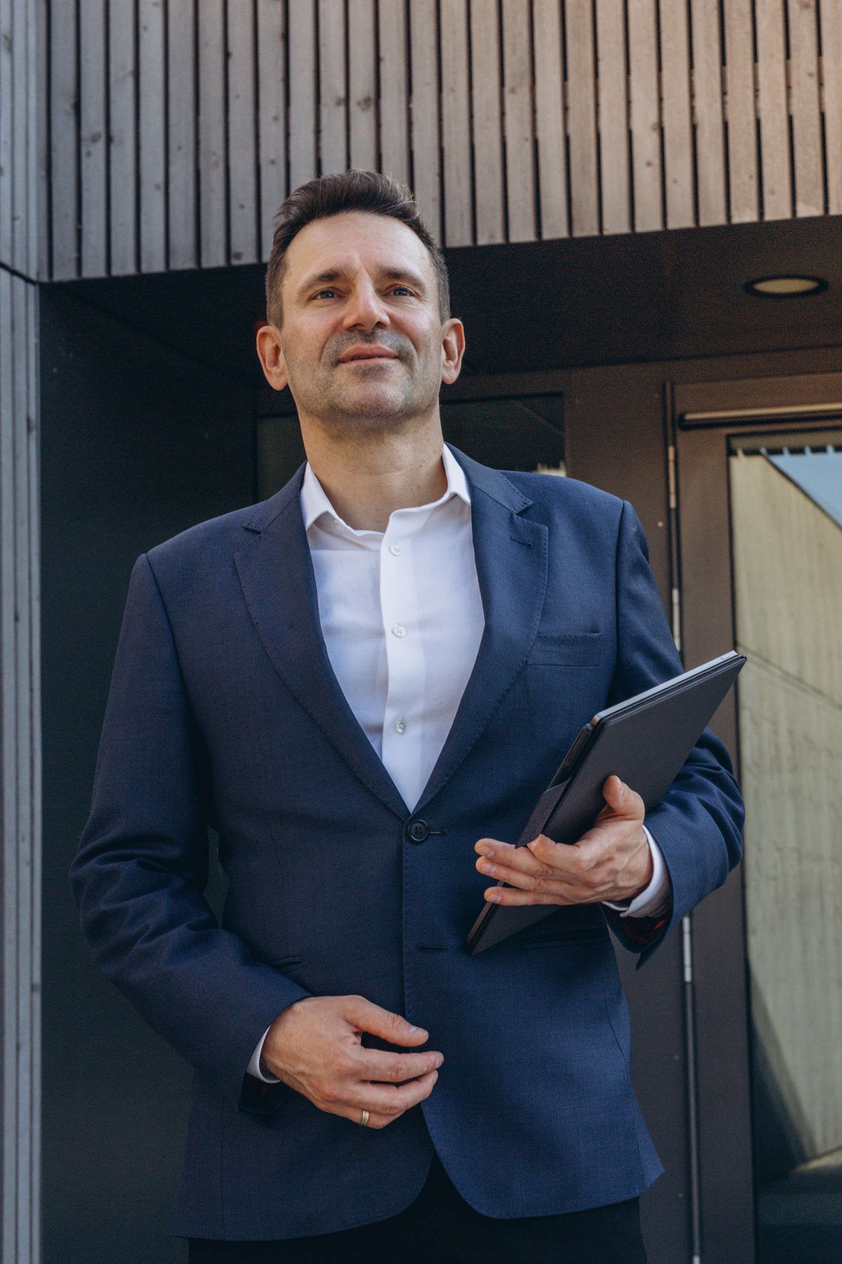 Frank Geiger im dunkelblauen Anzug mit Tablet vor einem modernen Gebäude