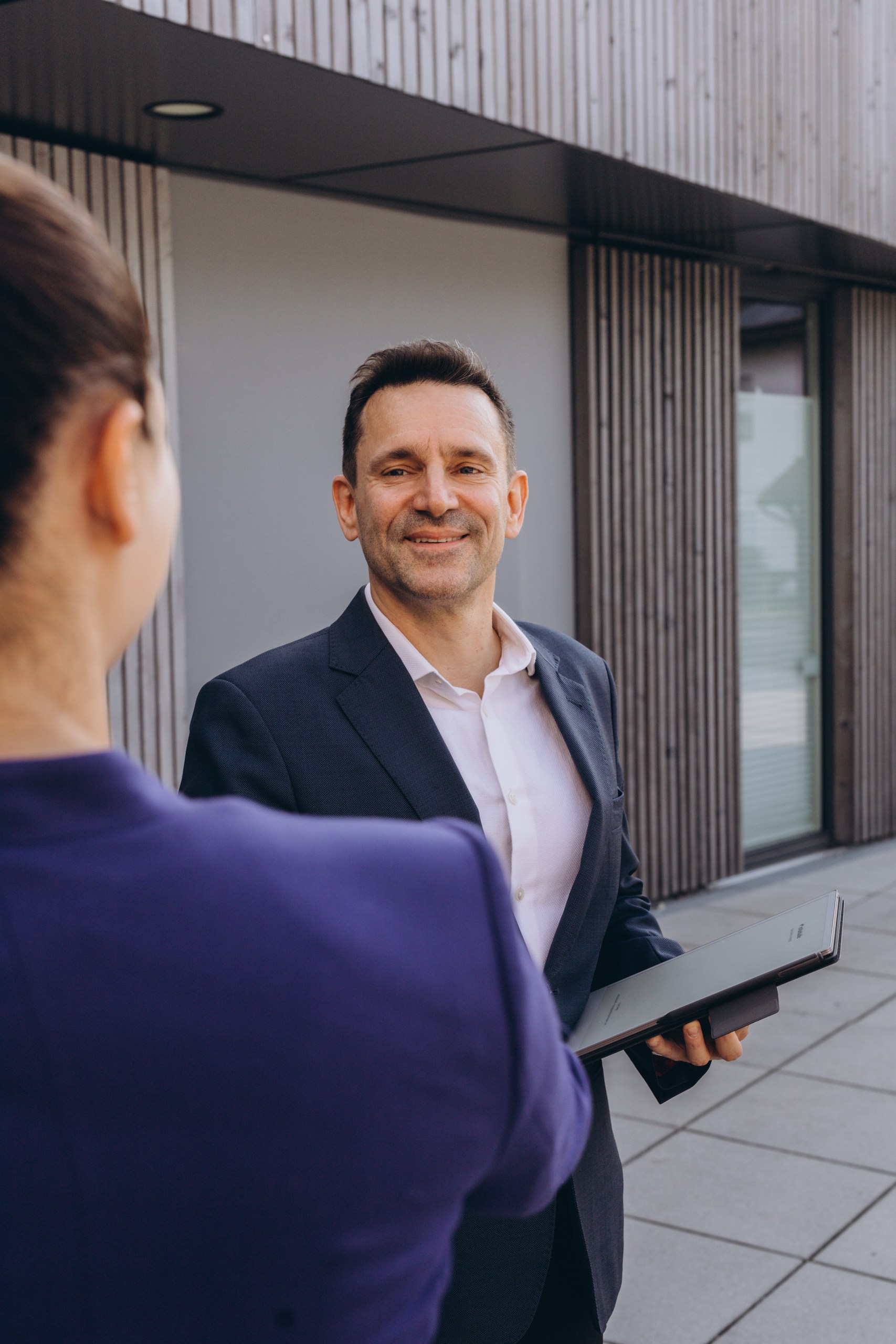Frank Geiger im Anzug mit Tablet im Gespräch vor einem modernen Gebäude